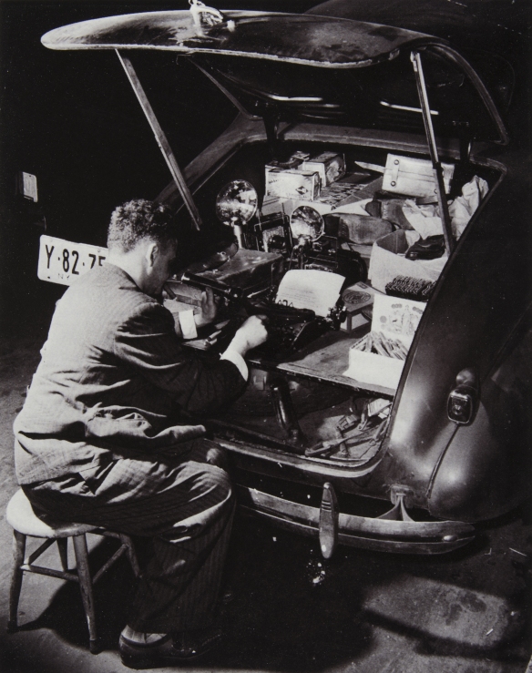 Weegee sentado en un taburete escribiendo a maquina en el maletero de su Chevrolet - Weegee International Center of Photography Cortesía Colección M. M. Auer